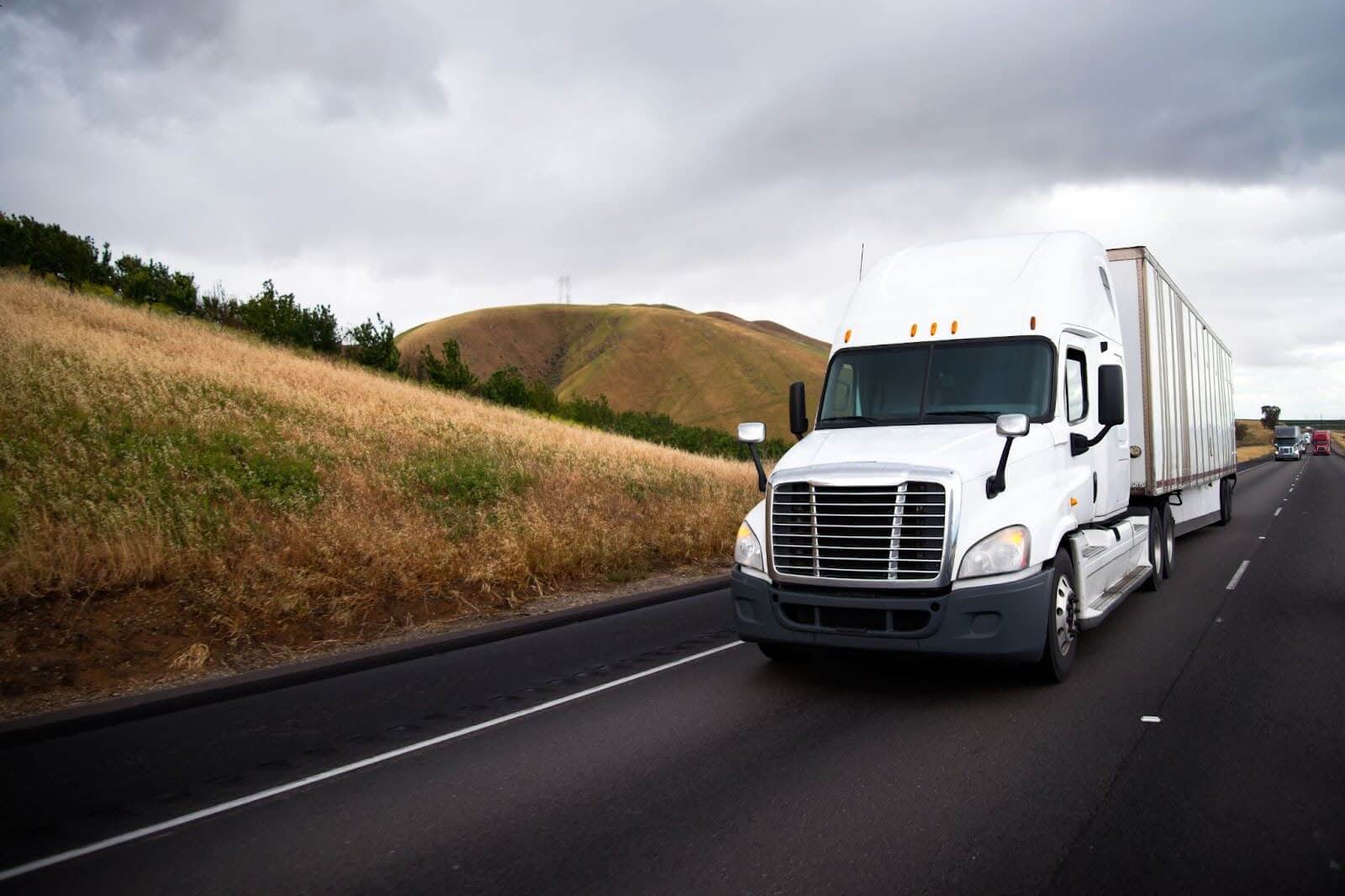 White big rig semi truck with dry van semi trailer on the highway with hill roadside in california